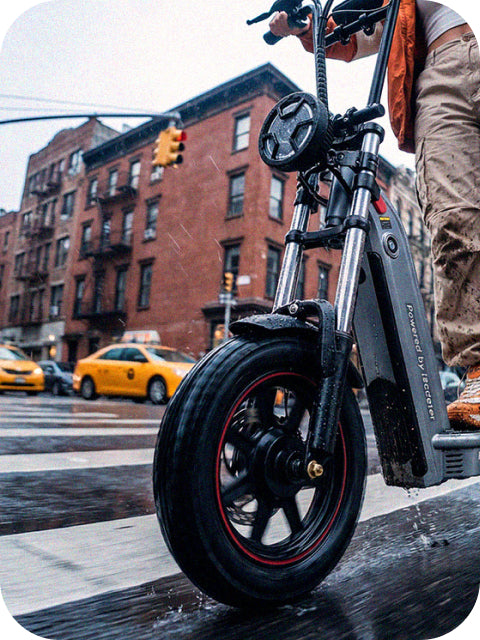 A close-up lifestyle photo focusing on the front wheel and sturdy chassis of an iScooter F7 Ultra electric scooter being ridden across a wet city crosswalk, with blurred yellow cabs and brick buildings in the background.