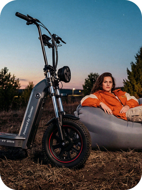 A lifestyle photo of an iScooter F7 Ultra electric scooter parked on a grassy field at dusk, with a young woman in an orange jacket relaxing on an inflatable lounge chair next to it.