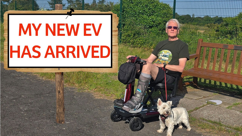 Thumbnail image showing a man seated on a mobility scooter with a small dog beside him next to a sign reading “MY NEW EV HAS ARRIVED,” outdoors near a park bench.