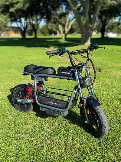 Front three-quarter view of a black iScooter DX5 electric mini-bike with a center basket and red rear shock absorbers parked on a grassy lawn under bright sunlight.