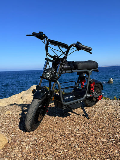 Black iScooter electric bike parked on a sunny, rocky coastal viewpoint overlooking the deep blue sea under a clear sky.