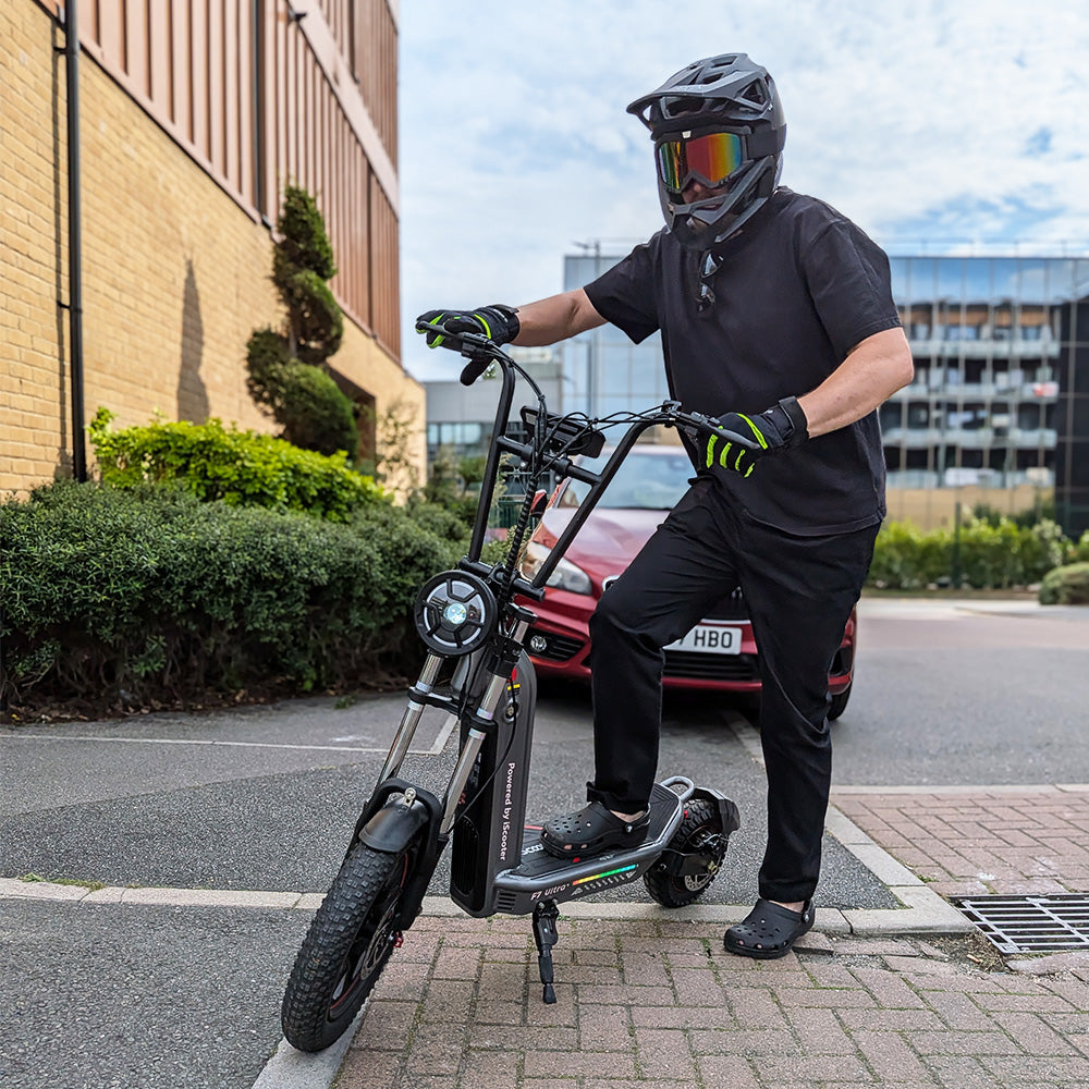 A rider wearing a full-face black helmet with colorful goggle lenses, black shirt, and gloves straddling a black iScooter F7 Ultra electric bike parked outdoors on a paved street.
