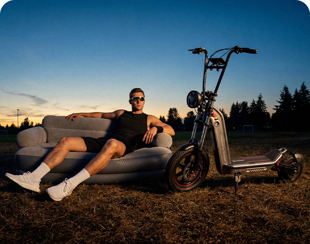 An outdoor lifestyle photo featuring a muscular man wearing a black tank top and sunglasses lounging on a grey inflatable sofa in a grassy field at dusk, with an iScooter F7 Ultra electric scooter parked upright right next to him under a fading twilight sky.