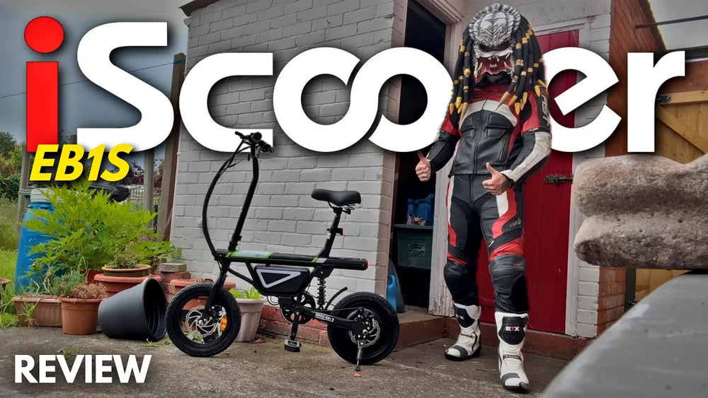 A video review thumbnail featuring a black iScooter EB1S electric bike and a person standing next to it outdoors wearing full motorcycle racing leathers and a stylized Predator-themed helmet.