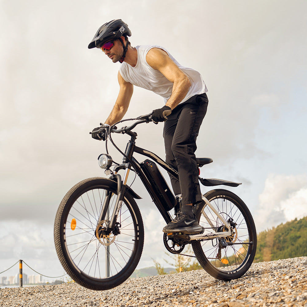 Person wearing a helmet and sunglasses riding a black electric bike on a rocky outdoor path with hills and clouds in the background.