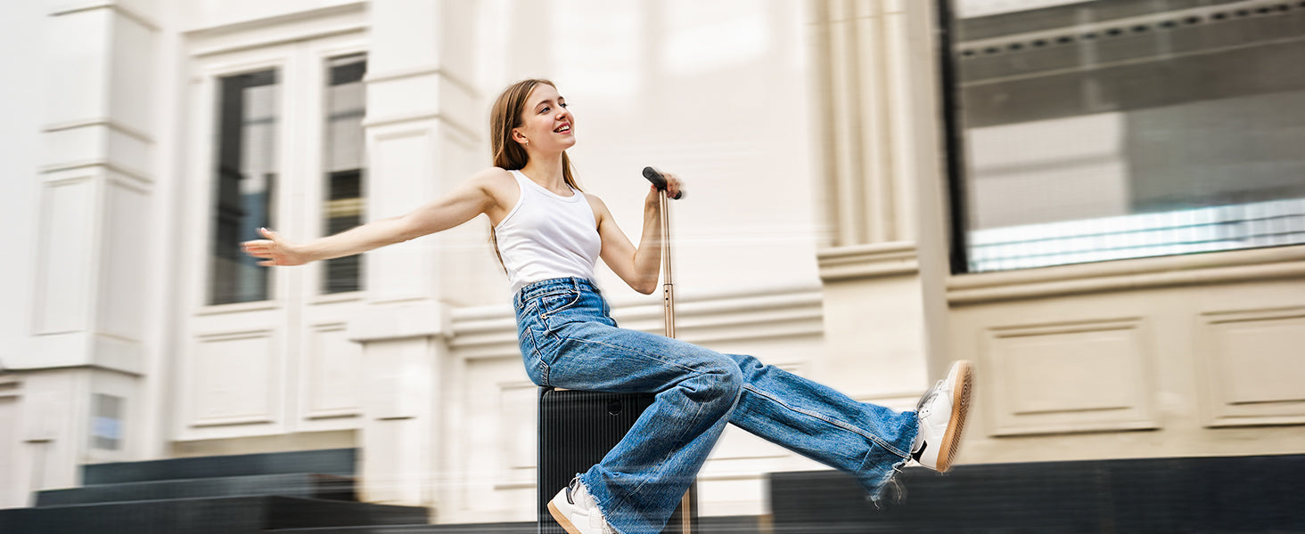 A wide product marketing banner featuring a joyful young woman riding a black PCD2501 rideable smart suitcase down a city street, captured with a motion blur effect. She is wearing a white tank top and blue jeans, smiling and holding the extended handlebar with one hand while stretching her other arm out to the side.