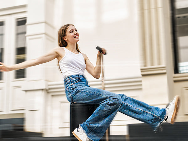 A joyful young woman riding a black PCD2501 rideable smart suitcase down a city street, captured with a motion blur effect. She is wearing a white tank top and blue jeans, smiling and holding the extended handlebar with one hand while extending her other arm out to the side.