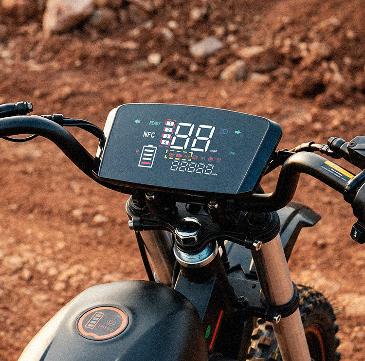 Close-up of the iScooter DX5 Max electric bike cockpit, featuring a large digital display mounted between the handlebars. The screen shows speed, battery level, riding mode, and status indicators, while the bike’s front suspension and frame are visible below. The rocky outdoor setting highlights the display’s visibility and functionality for off-road riding.