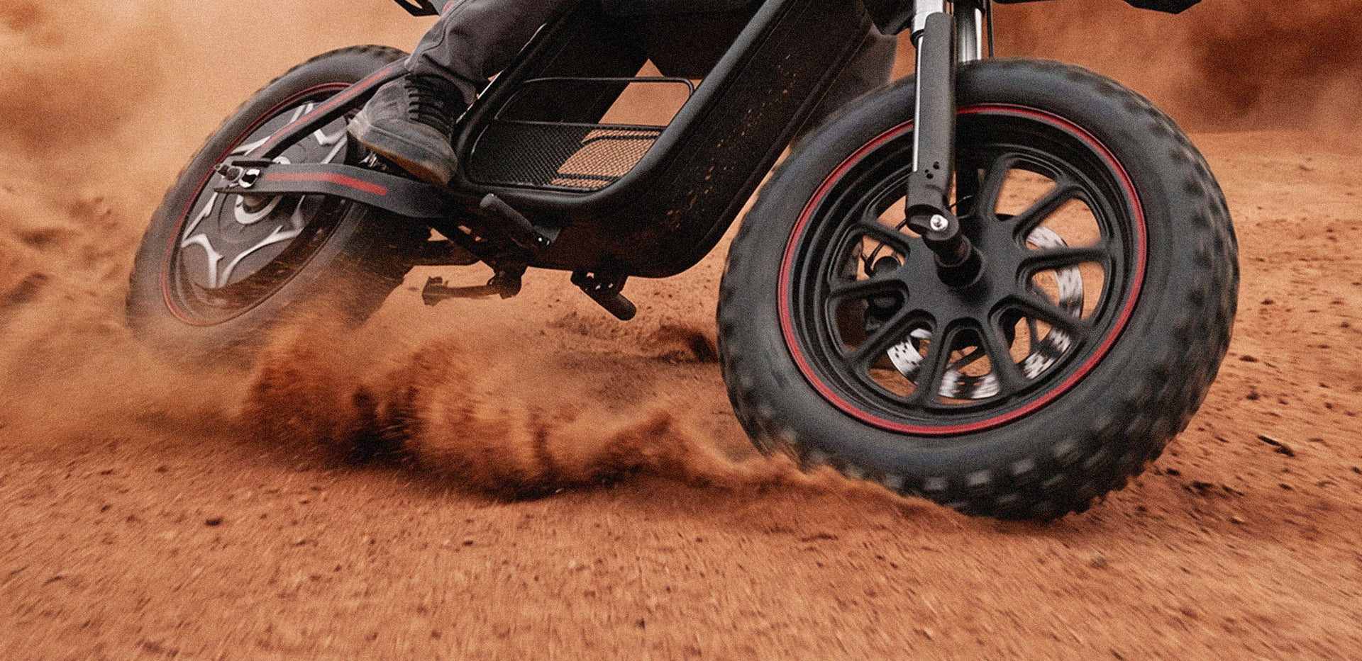 Wide-format close-up of the iScooter DX5 Max electric bike riding through sandy terrain, with its oversized fat tires prominently featured. Sand is thrown into the air as the tires grip the loose surface, highlighting traction, stability, and all-terrain performance. The panoramic composition emphasizes the bike’s rugged design and capability in challenging off-road conditions.