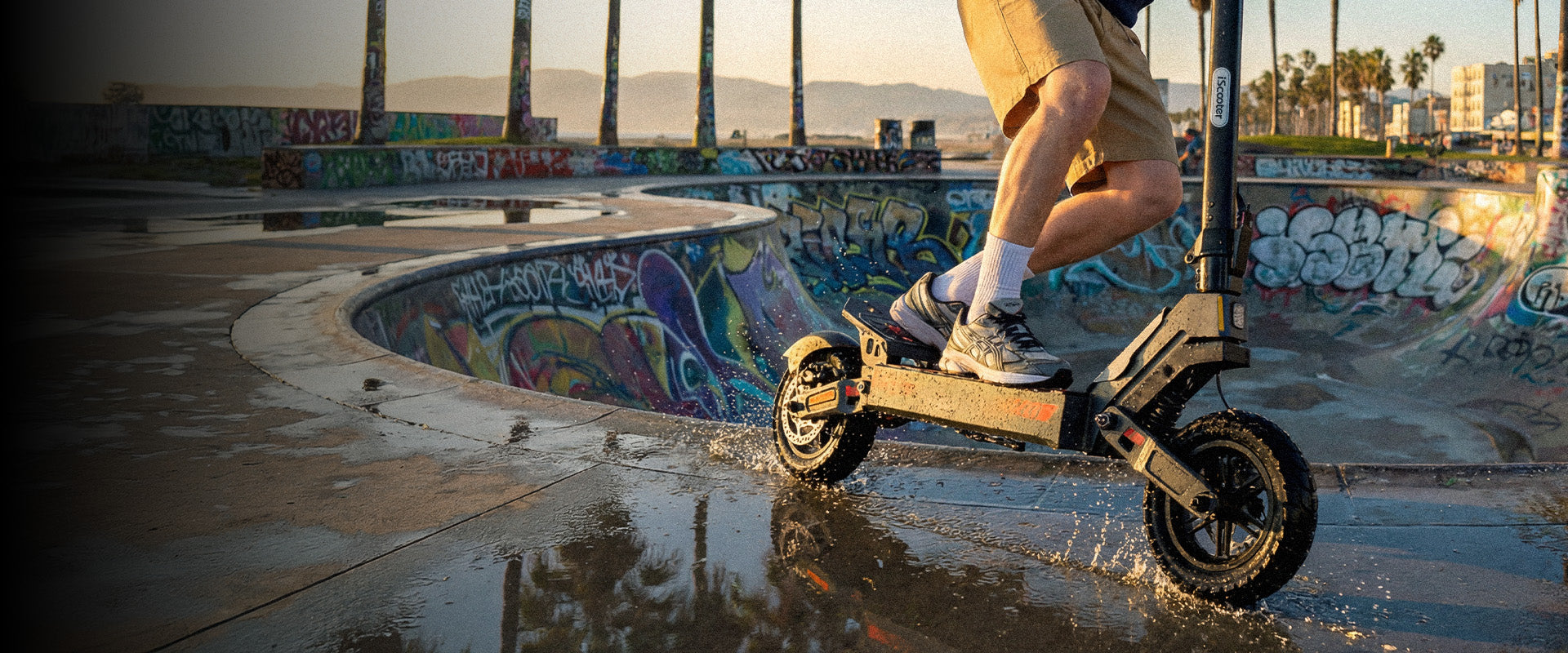 Person riding an electric scooter through a wet skatepark at sunset, splashing through puddles beside a graffiti-covered bowl, highlighting the scooter’s water-resistant performance and all-terrain capability.