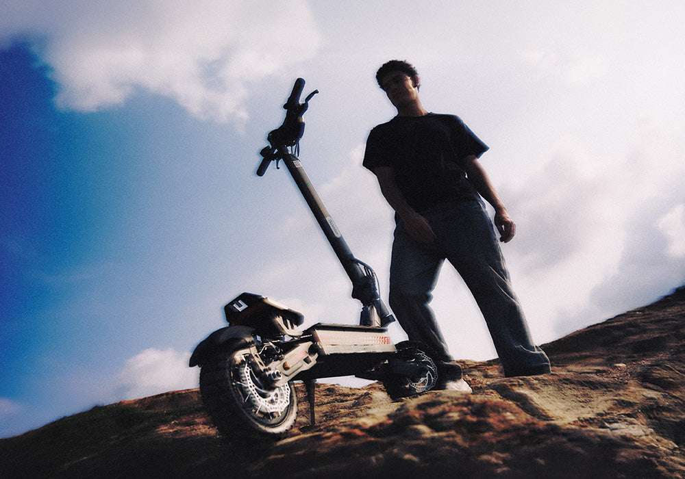 A person standing next to an electric scooter on a rocky hill against a cloudy sky.
