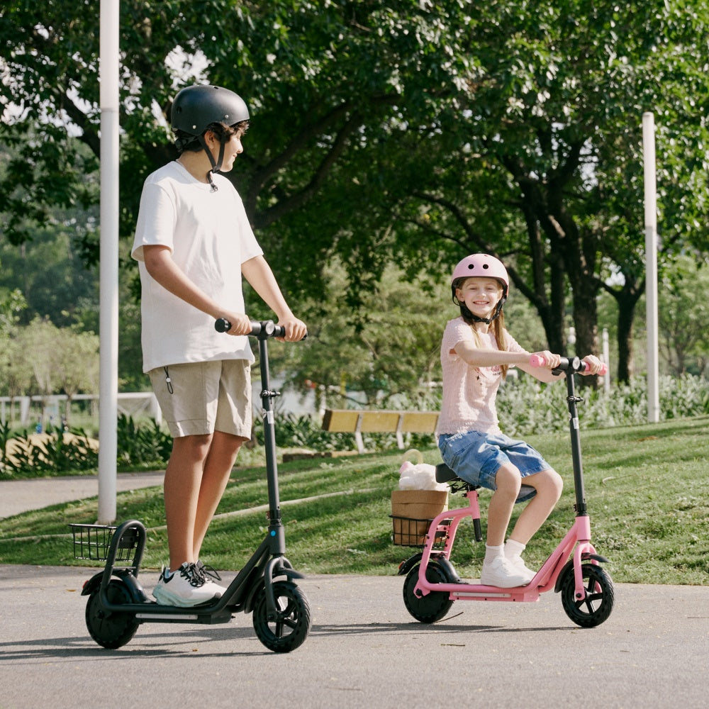 Two children wearing helmets ride electric scooters on a sunny park path, with the girl smiling on a pink seated scooter and the boy on a black scooter beside her.