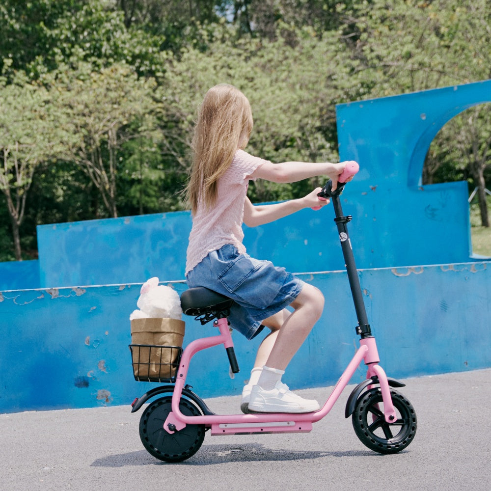 Girl riding a pink seated electric scooter with a rear storage basket on a paved path beside a blue skate park wall, wearing a pink helmet and enjoying an outdoor ride.