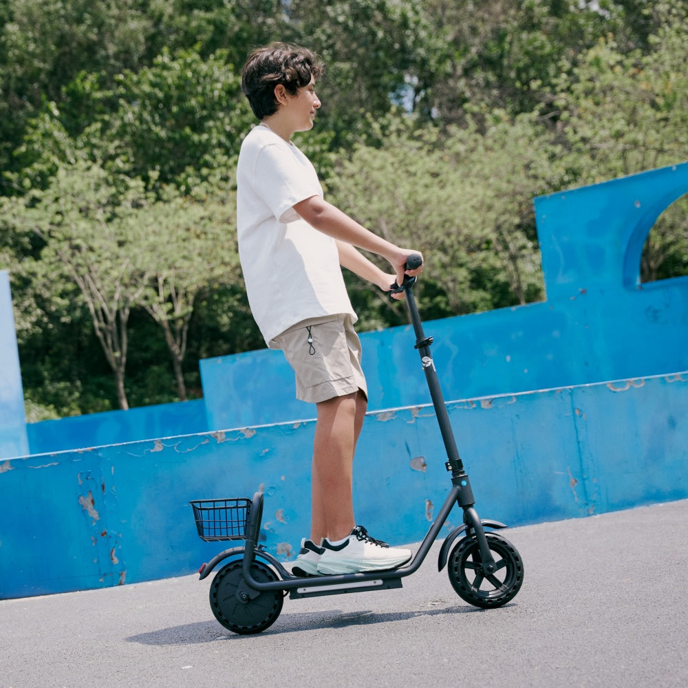 Child riding a black electric scooter with a rear basket along a paved path in a sunny park, viewed from the side.