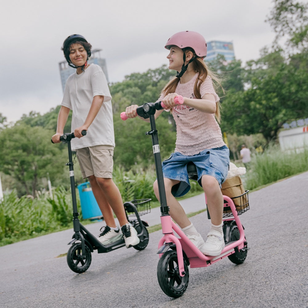 Two children wearing helmets ride electric scooters together on a paved park path, with trees and greenery in the background.