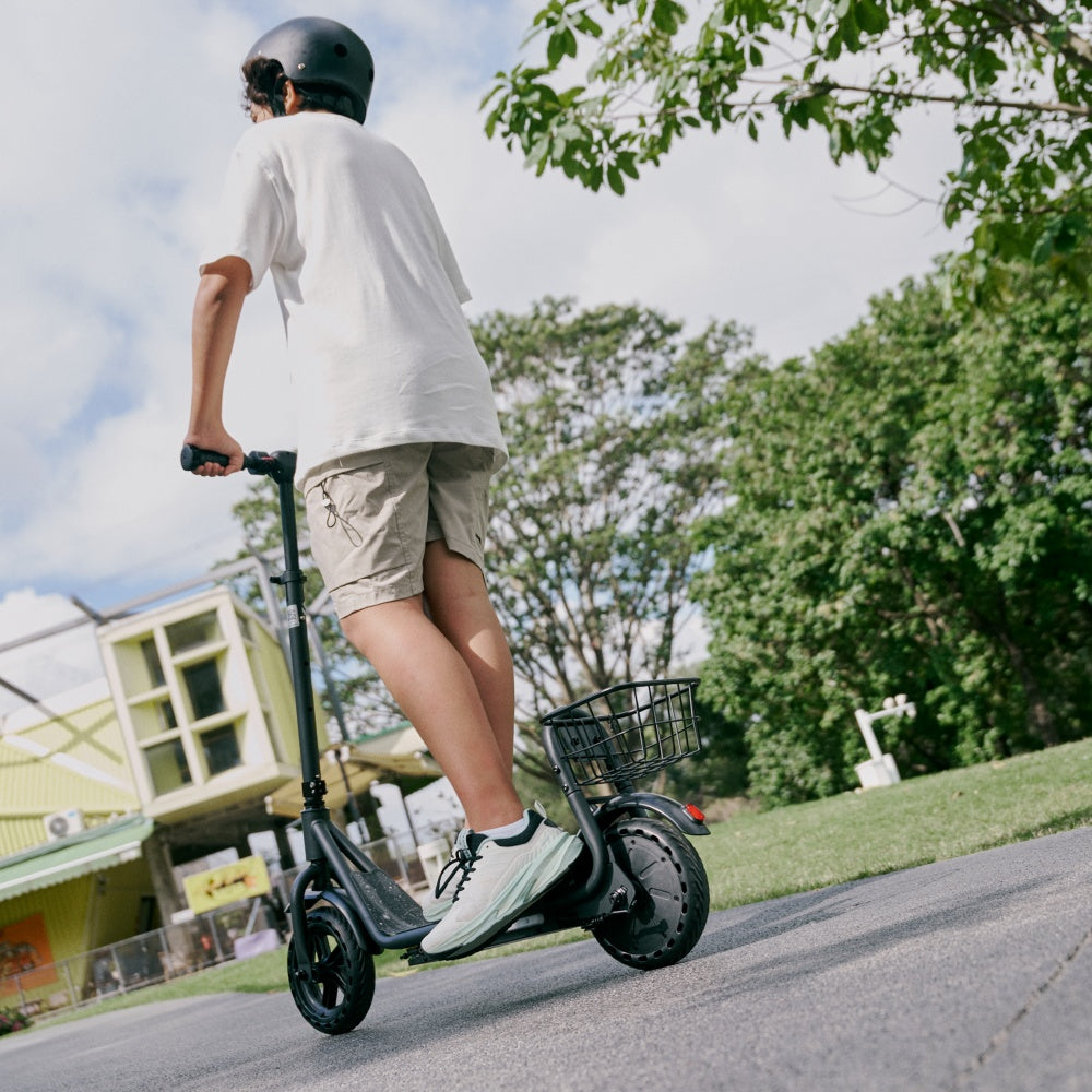 Boy riding a black electric scooter on a paved park path, viewed from behind, wearing a helmet with trees and blue sky in the background.