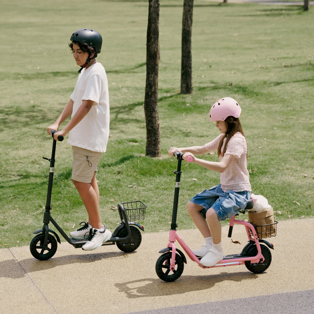 Two children wearing helmets ride electric scooters side by side on a sunny park pathway, with a boy on a black scooter and a girl on a pink seated scooter.