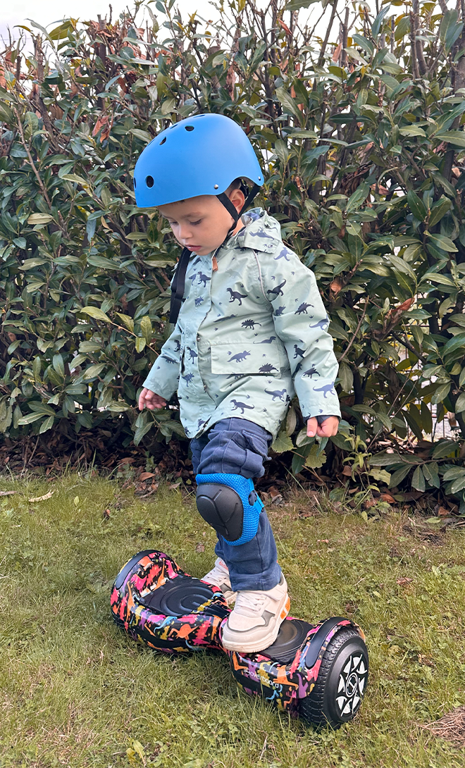Child wearing a helmet and riding a hoverboard on grass with bushes in the background