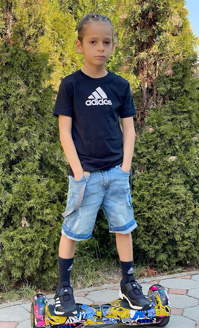 Child wearing an Adidas shirt standing on a hoverboard with greenery in the background