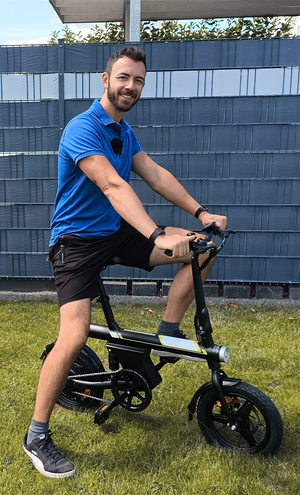Man riding a black electric bike on grass with a blue fence in the background