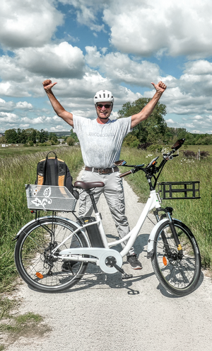 Person standing with arms raised next to a white electric bike in a field with a blue sky.