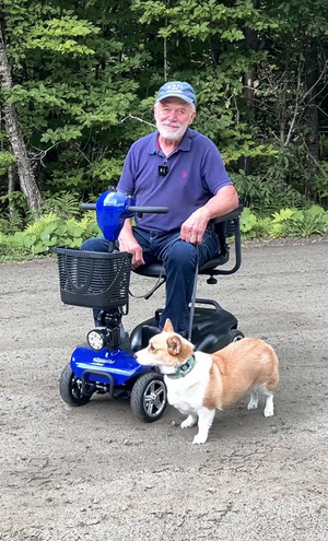 Man sitting on a mobility scooter with a dog standing next to him in a forested area.