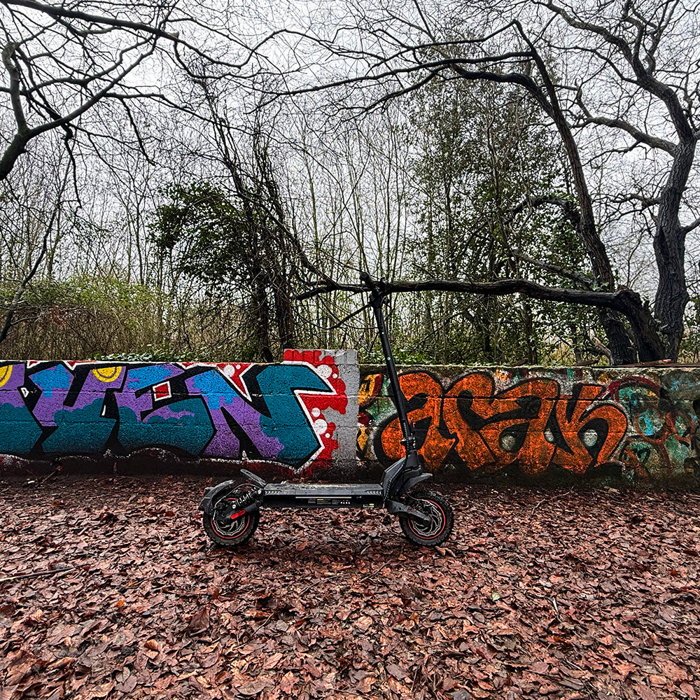 A black iScooter iX7 Pro electric scooter parked on fallen autumn leaves in front of a colorful graffiti wall.