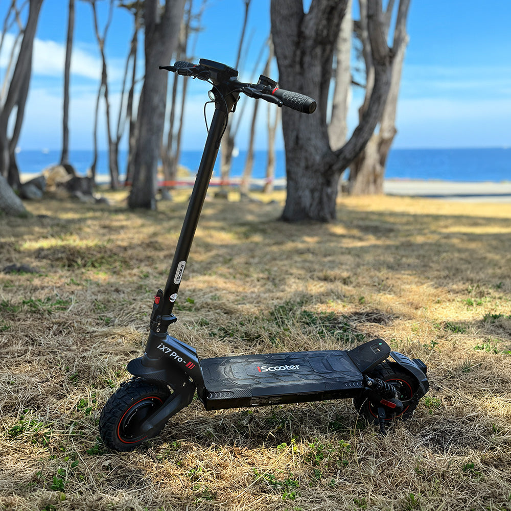 A black iScooter iX7 Pro electric scooter parked outdoors near a beach with trees and blue ocean in the background.