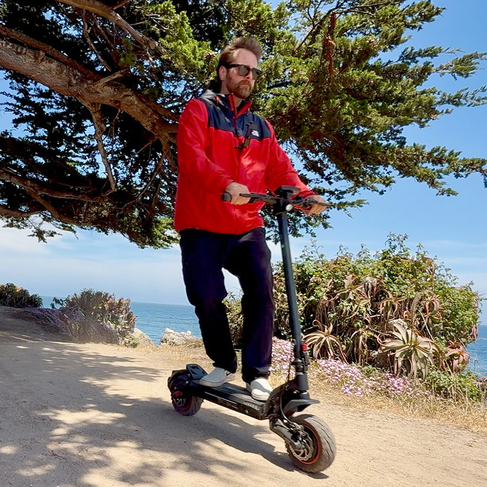 A man in a red jacket riding a black electric scooter along a sunny coastal path.