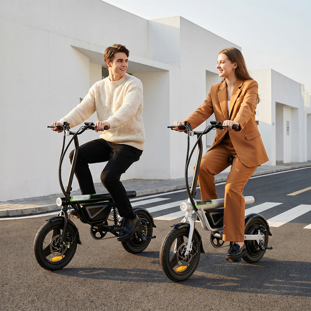 Two people riding compact electric bikes with thick tires and foldable frames along a clean urban street in the UK, smiling and looking at each other while cruising past modern white buildings.