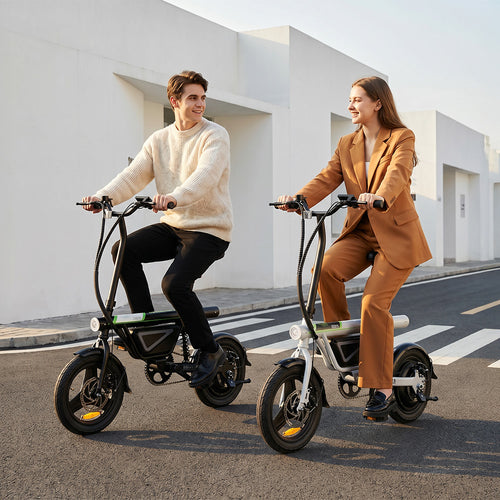 Two people riding compact electric bikes with thick tires and foldable frames along a clean urban street in the UK, smiling and looking at each other while cruising past modern white buildings.