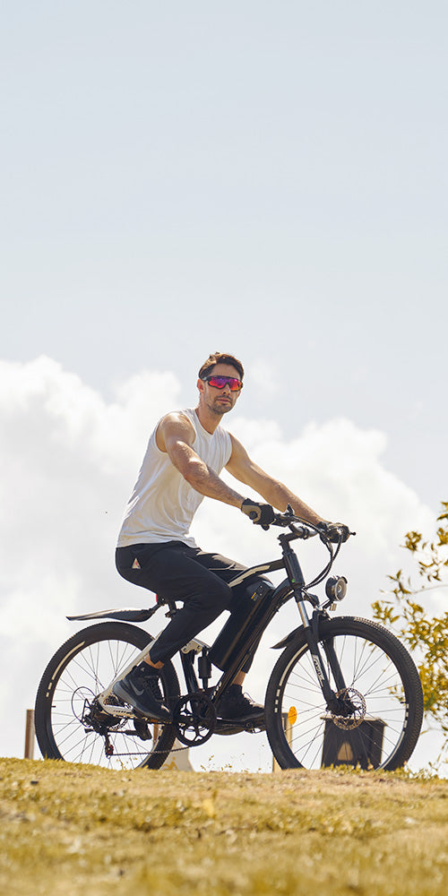 Person wearing sunglasses riding a black electric bike on a grassy hill under a bright sky with clouds in the background.