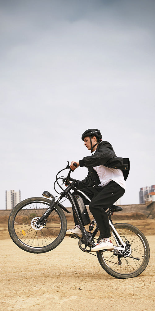 Person wearing a helmet performing a wheelie on a black M50 electric bike while riding on a dirt path outdoors under a cloudy sky.