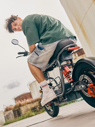 A low-angle rear view of a young man smiling over his shoulder while sitting on a parked grey DX5 electric scooter, highlighting the rear suspension and orange brake assembly.