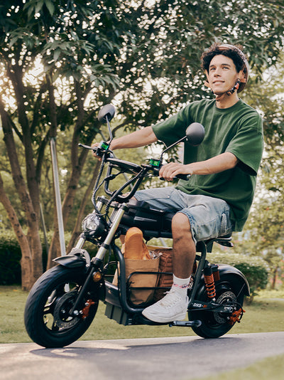 A young man wearing a helmet looks off-camera while riding a grey DX5 electric scooter down a paved path lined with sunlit green trees.