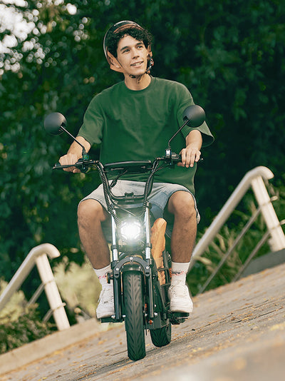 A young man wearing a helmet rides a grey DX5 electric scooter down a paved path, with a baguette tucked into the storage frame and lush green trees in the background.