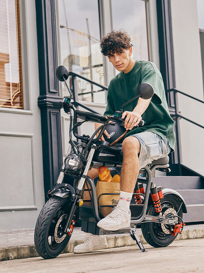 A young man holding a helmet sits on a parked grey DX5 electric scooter packed with a picnic basket, posing in front of a classic dark store facade.