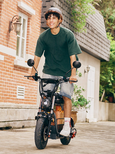 A young man wearing a helmet smiles while riding a grey DX5 electric scooter down a paved neighborhood street next to a brick building.