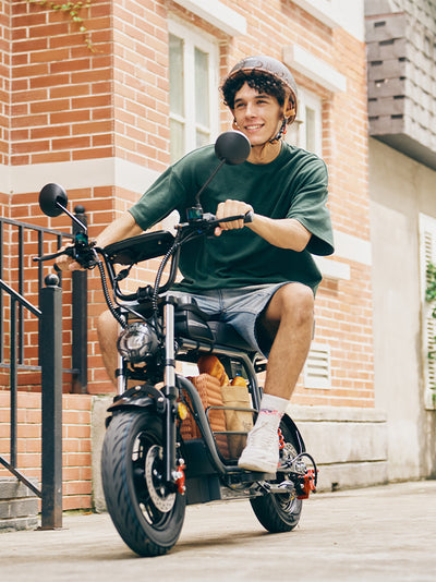 A young man sits casually on a grey DX5 electric scooter packed with a picnic basket, posing against a white architectural wall in bright sunlight.