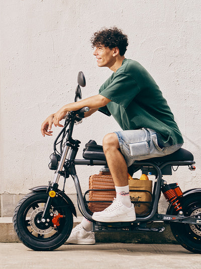 A side profile of a young man smiling while sitting on a parked grey DX5 electric scooter packed with a picnic basket against a textured white concrete wall.