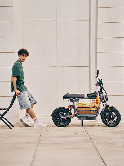 A young man leans on a folding chair next to a parked grey DX5 electric scooter, which is loaded with a picnic basket and a French baguette in its center storage frame.