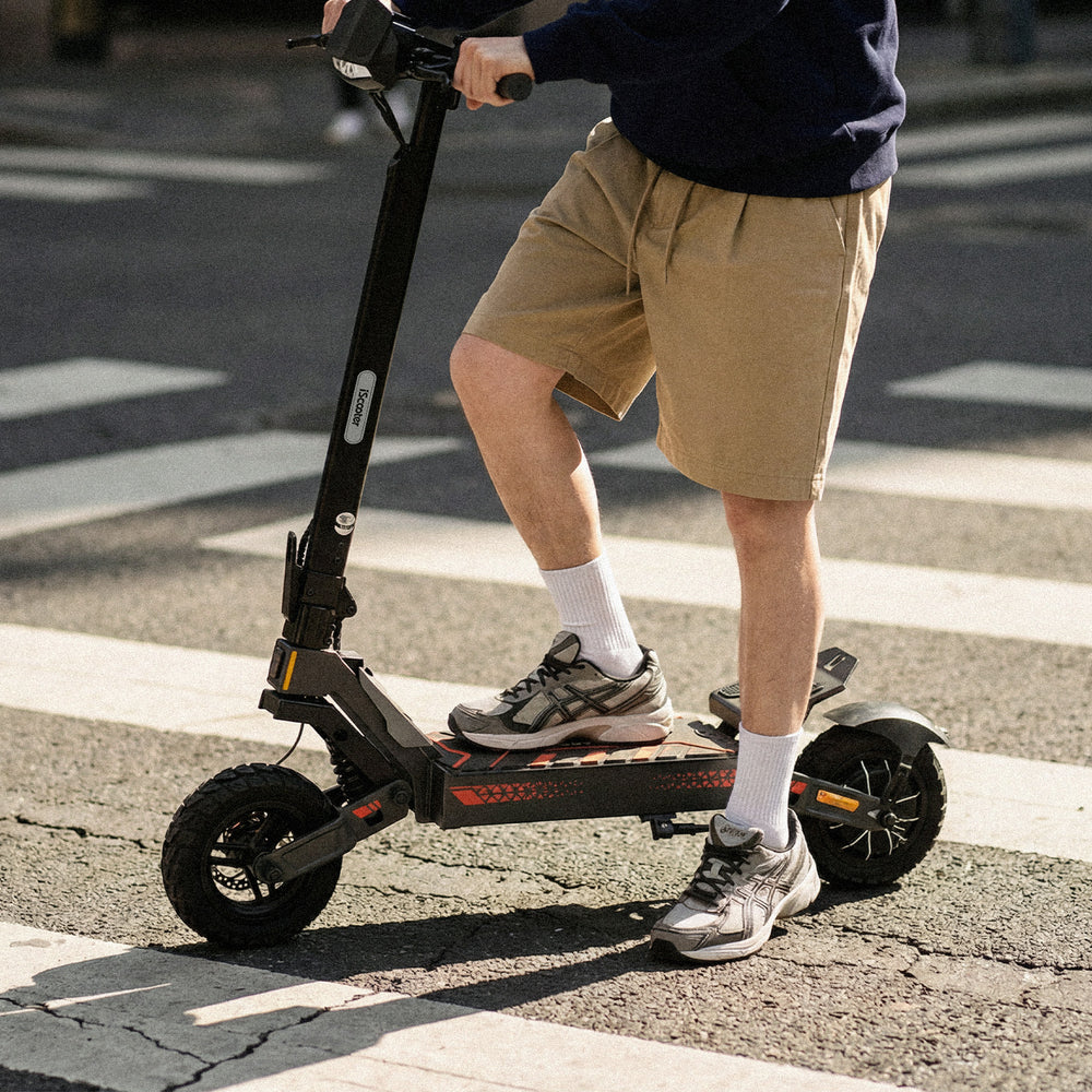 Person riding a black electric scooter across a city crosswalk. The image is tightly cropped to show the rider from the shoulders down, wearing a dark sweatshirt, tan shorts, white socks, and grey trainers. Sunlight casts shadows on the pavement, and the scooter has large tires and orange accents. The screenshot also shows a media management interface with image details and editing options displayed on the right side.