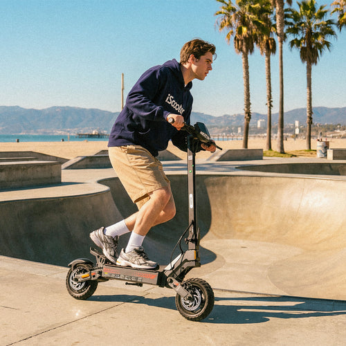 Young rider using an iScooter iF1 electric scooter at a sunny beachfront skate park, showcasing agile handling, sporty design, and urban outdoor riding with palm trees and ocean views in the background.