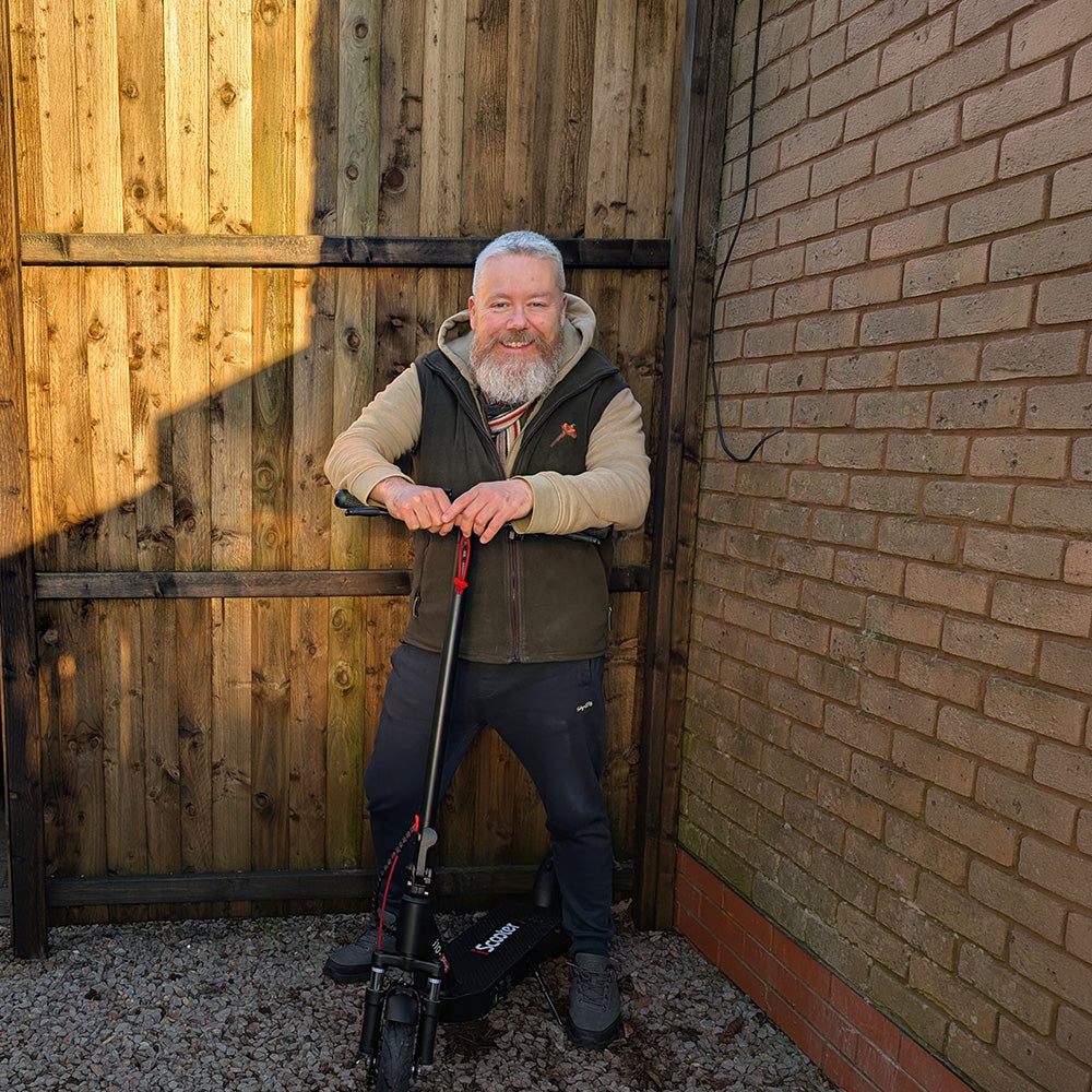 A smiling man with a beard standing with a black iScooter electric scooter against a wooden fence.