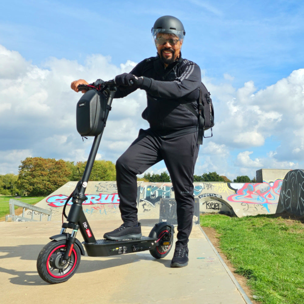 A man in black sportswear and a helmet standing with one foot on a black iScooter i10 Plus electric scooter at a skatepark.