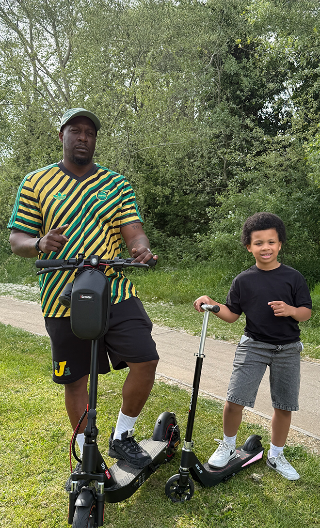 An outdoor lifestyle photo featuring a man in a green and yellow striped jersey standing next to an iScooter i10Max electric scooter, and a young boy standing beside an iHoverboard iK1s electric scooter on a grassy path with trees in the background.
