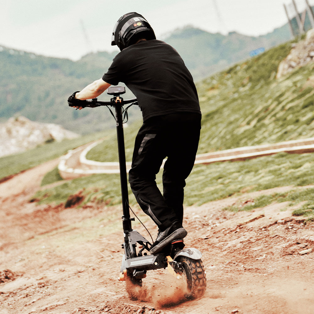 Rear view of a rider in full gear riding an off-road electric scooter downhill on a dirt path.