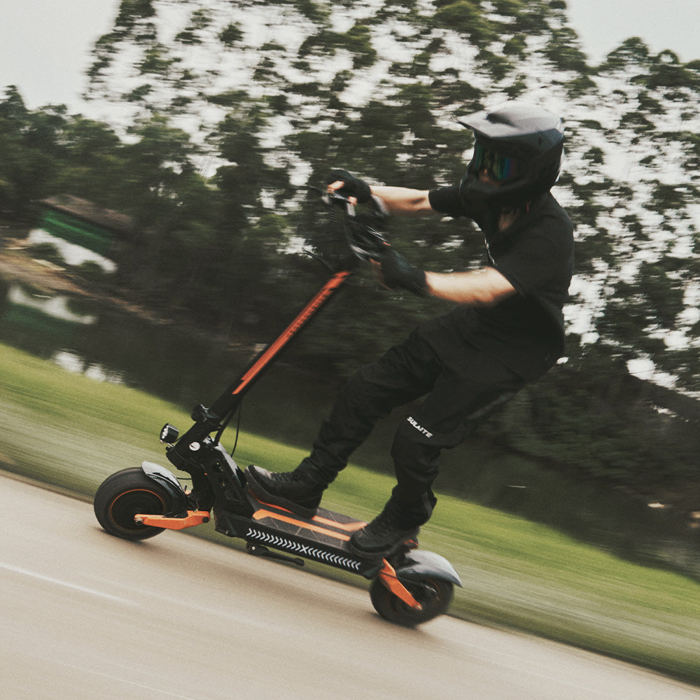 A rider in full gear riding an off-road electric scooter fast along a paved park path.