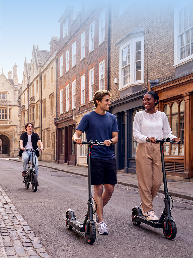 Two adults riding iScooter electric scooters through a historic city street with an e-bike rider behind, showcasing everyday urban commuting.