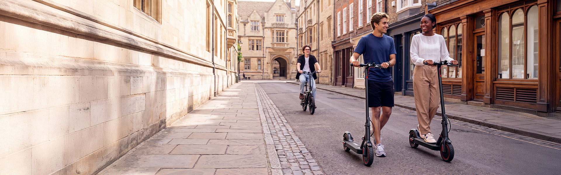Two adults riding iScooter electric scooters through a historic city street with an e-bike rider behind, highlighting urban commuting.